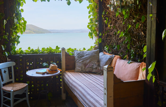 The romantic sea view from the Angel’s Lair balcony at TwoAngels in Knysna. In the background is a sunny sky, mountains and blue water. In the foreground is a comfortable looking daybed with lots of pillows. The green leaves of a Bougainvillea surround the balcony and frame the view.